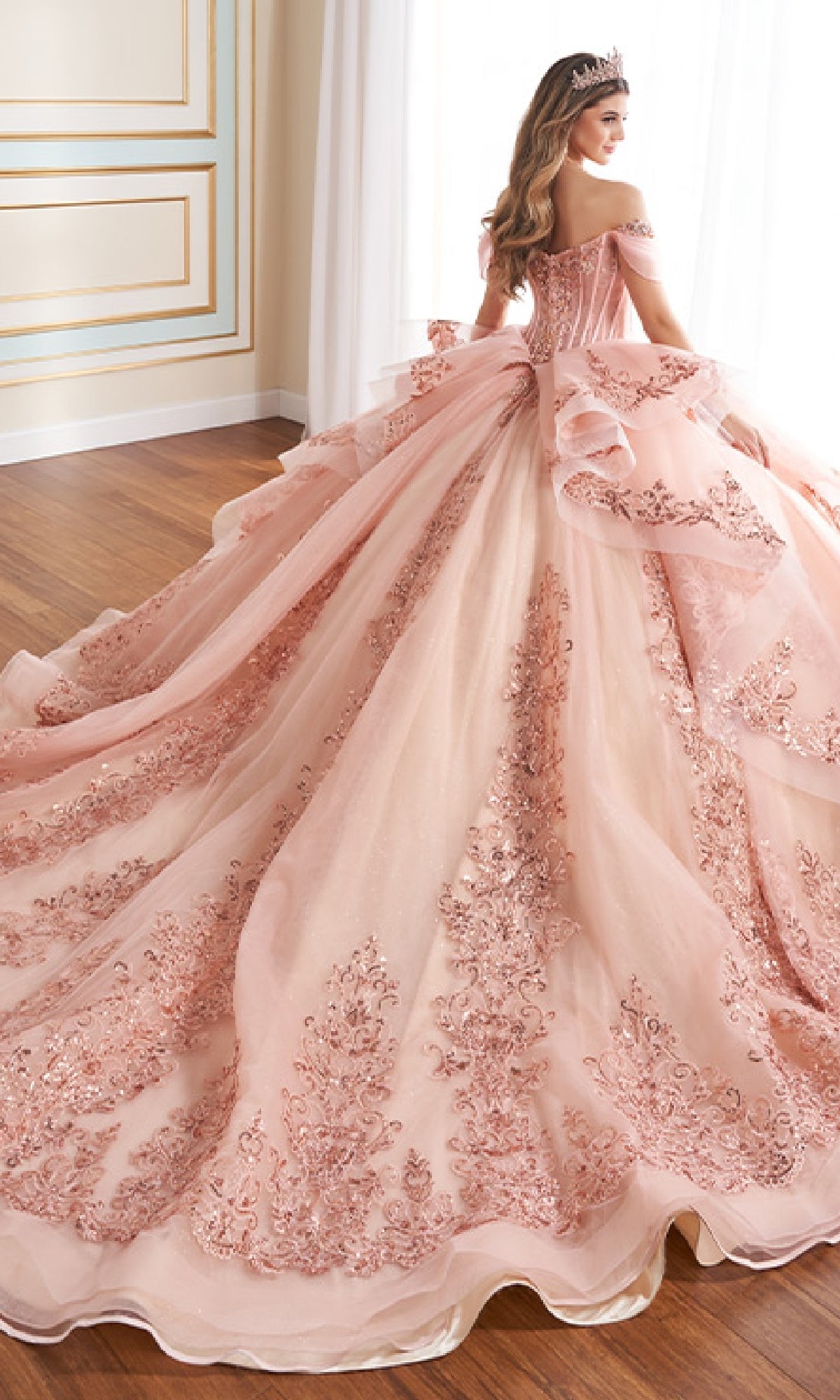A woman stands indoors near a window wearing the Quinceanera Dress PR30171 By Princesa, a pink off-the-shoulder gown with intricate embroidery, a voluminous glitter tulle train, and a tiara.